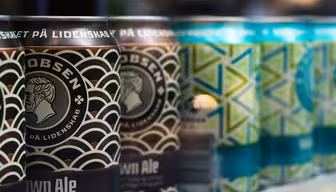 Close-up of beer cans in two rows. The foreground shows dark brown cans with an intricate pattern and a logo featuring a profile of a man. The background has teal and green cans with geometric designs. The text is mostly in Danish.