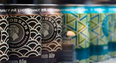 Close-up of beer cans in two rows. The foreground shows dark brown cans with an intricate pattern and a logo featuring a profile of a man. The background has teal and green cans with geometric designs. The text is mostly in Danish.