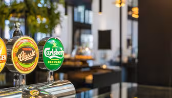 Close-up of a bar with beer taps featuring Tuborg Classic and Carlsberg logos. The background has a modern interior with plants and soft lighting.