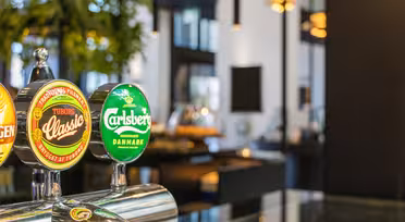 Close-up of a bar with beer taps featuring Tuborg Classic and Carlsberg logos. The background has a modern interior with plants and soft lighting.