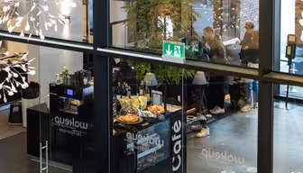 View from above of a modern café with glass walls, showcasing customers ordering at the counter. Pastries and drinks are displayed. People are visible through the glass entrance, and a green exit sign hangs on the ceiling.
