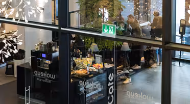 View from above of a modern café with glass walls, showcasing customers ordering at the counter. Pastries and drinks are displayed. People are visible through the glass entrance, and a green exit sign hangs on the ceiling.