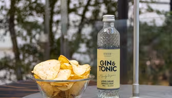 A clear glass bowl filled with potato chips sits next to a bottle of ready-to-serve gin and tonic on an outdoor table. The background features blurred greenery, suggesting a relaxed, natural setting.