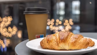 A flaky croissant sits on a white plate beside a brown takeaway coffee cup with a black lid. The background features soft, blurred lights, creating a cozy and inviting atmosphere.