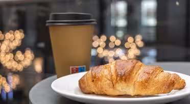 A flaky croissant sits on a white plate beside a brown takeaway coffee cup with a black lid. The background features soft, blurred lights, creating a cozy and inviting atmosphere.