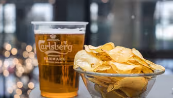 A clear plastic cup of Carlsberg beer stands next to a glass bowl filled with potato chips on a table. The background features blurred lights, creating a festive atmosphere.