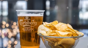 A clear plastic cup of Carlsberg beer stands next to a glass bowl filled with potato chips on a table. The background features blurred lights, creating a festive atmosphere.