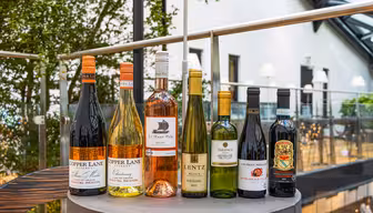 A selection of eight wine bottles on a round table in a modern indoor setting. The bottles vary in colors and labels, with some marked Copper Lane and Lentz. Glass walls and greenery are visible in the background.