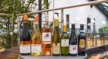 A selection of eight wine bottles on a round table in a modern indoor setting. The bottles vary in colors and labels, with some marked Copper Lane and Lentz. Glass walls and greenery are visible in the background.
