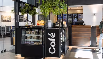 Modern café kiosk with glass display of pastries and snacks. Black counter with Café written vertically. Potted plants suspended above, and fridges with drinks visible in the background. Bright lighting and sleek design.