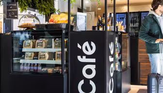 A café display featuring snacks, pastries, and packaged goods. A glass cover showcases croissants with a coffee and croissant deal sign. A person with luggage is partially visible on the right. Greenery hangs from above, adding a touch of nature.