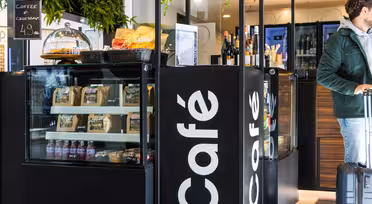 A café display featuring snacks, pastries, and packaged goods. A glass cover showcases croissants with a coffee and croissant deal sign. A person with luggage is partially visible on the right. Greenery hangs from above, adding a touch of nature.