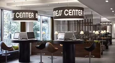 Spacious modern hotel lobby with a business center. Features computer stations on round tables with barstool seating. Large overhead lights display the text Business Center. The space is well-lit with views of trees outside.