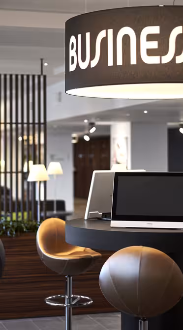 Modern business center with sleek desks, computers on round tables, and stylish chairs. Large hanging lights display the text Business Center. Background features contemporary decor and natural lighting.