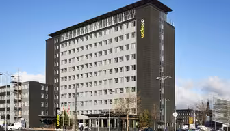 A modern multi-story hotel building with a dark facade and rows of windows. The name Wakeup is visible on the building. Several cars are parked nearby, and a clear blue sky is in the background.