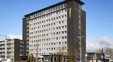 A modern multi-story hotel building with a dark facade and rows of windows. The name Wakeup is visible on the building. Several cars are parked nearby, and a clear blue sky is in the background.