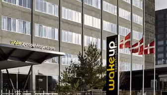 The image shows the exterior of Wakeup Copenhagen hotel with its modern facade. Danish flags are seen flying nearby, and there is a sign with the hotels logo and website.