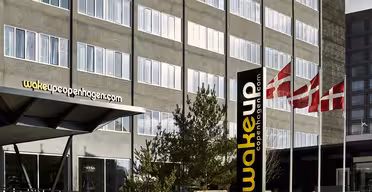 The image shows the exterior of Wakeup Copenhagen hotel with its modern facade. Danish flags are seen flying nearby, and there is a sign with the hotels logo and website.
