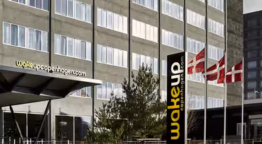 The image shows the exterior of Wakeup Copenhagen hotel with its modern facade. Danish flags are seen flying nearby, and there is a sign with the hotels logo and website.