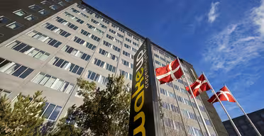 A tall building with numerous windows rises under a blue sky. Four red flags with white crosses on flagpoles are in front. A black and yellow sign near the entrance reads Wakeup Copenhagen. Trees are visible near the base of the building.