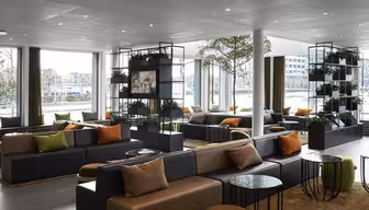 Modern lounge area with large windows, black and gray sofas, and colorful cushions. Potted plants decorate the room, and various round tables are placed around the seating. Natural light fills the space, creating a cozy atmosphere.