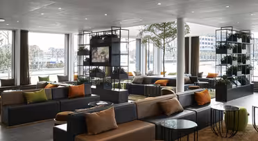 Modern lounge area with large windows, black and gray sofas, and colorful cushions. Potted plants decorate the room, and various round tables are placed around the seating. Natural light fills the space, creating a cozy atmosphere.