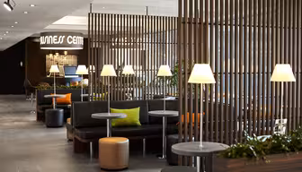 A modern relaxation area with partitioned wooden slats, cozy black seating, small round tables, and soft lighting from table lamps. Bright green and orange cushions add color, and potted plants enhance the serene atmosphere.