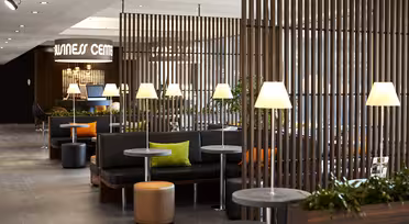 A modern relaxation area with partitioned wooden slats, cozy black seating, small round tables, and soft lighting from table lamps. Bright green and orange cushions add color, and potted plants enhance the serene atmosphere.