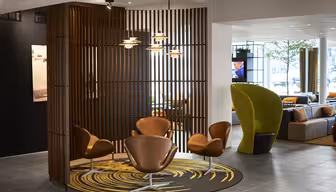 A modern seating area with four brown chairs around a circular rug featuring a yellow pattern. The space is partially enclosed by vertical wooden slats. Pendant lights hang above, and a separate area with green chairs is visible in the background.