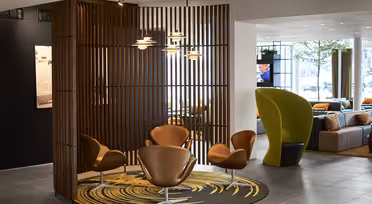 A modern seating area with four brown chairs around a circular rug featuring a yellow pattern. The space is partially enclosed by vertical wooden slats. Pendant lights hang above, and a separate area with green chairs is visible in the background.