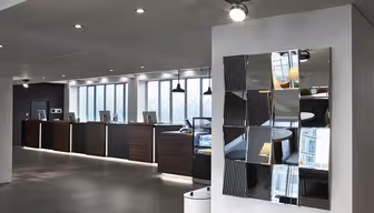 Modern hotel reception area with a sleek design. Dark wood counters lined with computers are set against large windows. A wall features a geometric mirrored artwork. Bright overhead lights illuminate the space, adding a contemporary feel.