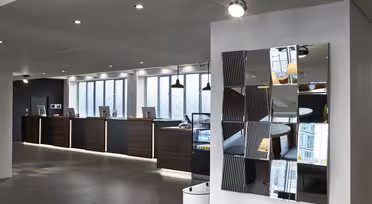 Modern hotel reception area with a sleek design. Dark wood counters lined with computers are set against large windows. A wall features a geometric mirrored artwork. Bright overhead lights illuminate the space, adding a contemporary feel.