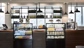 A modern café with wooden counters and hanging pendant lights. Glass display cases showcase beverages, snacks, and prepared foods. Shelves above hold wine bottles and plants, with a bright window in the background.