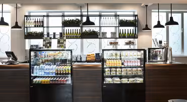 A modern café with wooden counters and hanging pendant lights. Glass display cases showcase beverages, snacks, and prepared foods. Shelves above hold wine bottles and plants, with a bright window in the background.