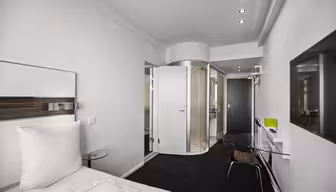 Modern hotel room with a white bed on the left, a glass desk and chair in the center, and a curved glass shower visible through an open bathroom door. The room has white walls, a black carpet, and recessed lighting.