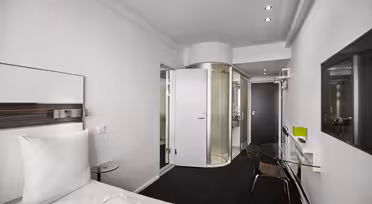 Modern hotel room with a white bed on the left, a glass desk and chair in the center, and a curved glass shower visible through an open bathroom door. The room has white walls, a black carpet, and recessed lighting.