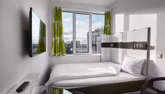 Small modern bedroom with a single bed, white bedding, a wall-mounted TV, and a glass desk. Green and white patterned curtains frame a window showing a cityscape.