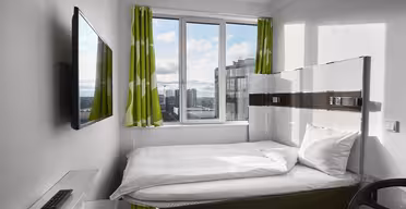 Small modern bedroom with a single bed, white bedding, a wall-mounted TV, and a glass desk. Green and white patterned curtains frame a window showing a cityscape.