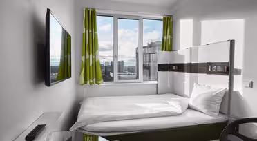 Small modern bedroom with a single bed, white bedding, a wall-mounted TV, and a glass desk. Green and white patterned curtains frame a window showing a cityscape.