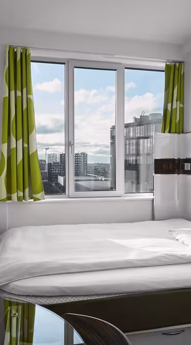 Small modern bedroom with a single bed, white bedding, a wall-mounted TV, and a glass desk. Green and white patterned curtains frame a window showing a cityscape.