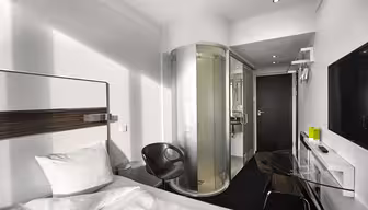 Modern hotel room with a white bed, a round glass-enclosed shower, a black chair, a glass desk with a black lamp, and a flat-screen TV on the wall. The design is sleek with a minimalist aesthetic.