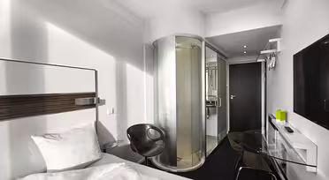 Modern hotel room with a white bed, a round glass-enclosed shower, a black chair, a glass desk with a black lamp, and a flat-screen TV on the wall. The design is sleek with a minimalist aesthetic.