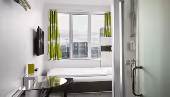 Small, modern hotel room with a neatly made bed, green and white curtains, wall-mounted TV, and a glass desk with a black chair. A tall window offers a cityscape view. Glass shower door and toiletries in the foreground.