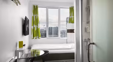 Small, modern hotel room with a neatly made bed, green and white curtains, wall-mounted TV, and a glass desk with a black chair. A tall window offers a cityscape view. Glass shower door and toiletries in the foreground.