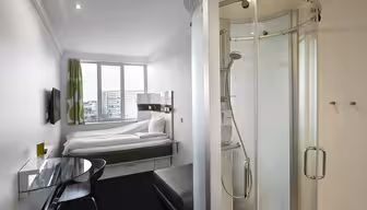 Compact modern hotel room with white walls and a large window. Features a bed, wall-mounted TV, small desk, and chair. A transparent shower stall is in the corner. Green curtains add a touch of color.