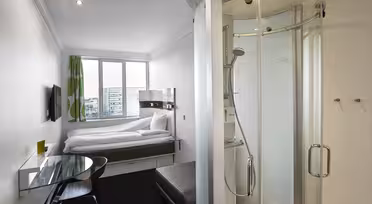 Compact modern hotel room with white walls and a large window. Features a bed, wall-mounted TV, small desk, and chair. A transparent shower stall is in the corner. Green curtains add a touch of color.