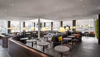 Modern dining area with round tables and wooden chairs. Each table is lit by small lamps. Large windows provide natural light, and a wall features green plants. The space is open and contemporary with a neutral color palette.