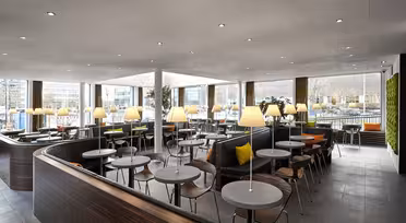 Modern dining area with round tables and wooden chairs. Each table is lit by small lamps. Large windows provide natural light, and a wall features green plants. The space is open and contemporary with a neutral color palette.