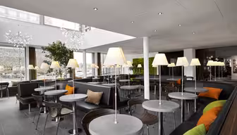Modern cafe interior with round tables and black seating, accented by orange and green cushions. Tall lamps are placed on most tables. Large windows bring in natural light, and greenery is part of the decor. White ceiling with recessed lights.