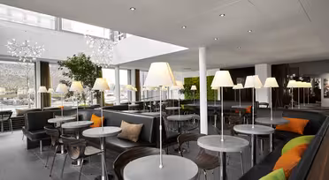 Modern cafe interior with round tables and black seating, accented by orange and green cushions. Tall lamps are placed on most tables. Large windows bring in natural light, and greenery is part of the decor. White ceiling with recessed lights.